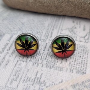 May include: A pair of silver stud earrings with a round, green, yellow, and red design featuring a black marijuana leaf.