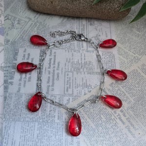 Peut inclure: Un bracelet en chaîne argenté orné de sept breloques rouges translucides en forme de goutte. Les breloques sont espacées uniformément le long de la chaîne, créant un design délicat et attrayant. Le bracelet est présenté sur une surface à motifs.