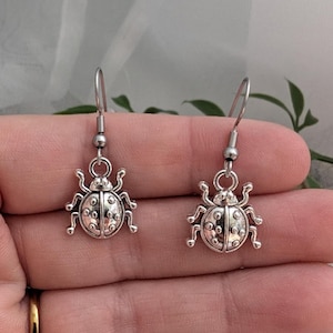 May include: A pair of silver-toned metal earrings featuring a ladybird charm design. Each earring has a dangling ladybird pendant with raised circular details on the wings and a simple hook-style ear wire for attachment to the earlobe.