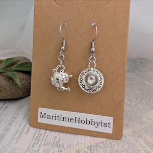 May include: Silver-coloured teapot and teacup dangle earrings. The earrings feature a small teapot charm and a teacup charm with a clear gem in the centre. The earrings are displayed on a brown card with the text "Maritime Hobbyist".