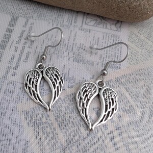 May include: A pair of silver-colored earrings featuring a wing design. Each earring has a hook for pierced ears and a detailed feather pattern within the wing shape. The earrings are displayed against a textured, light-colored background.