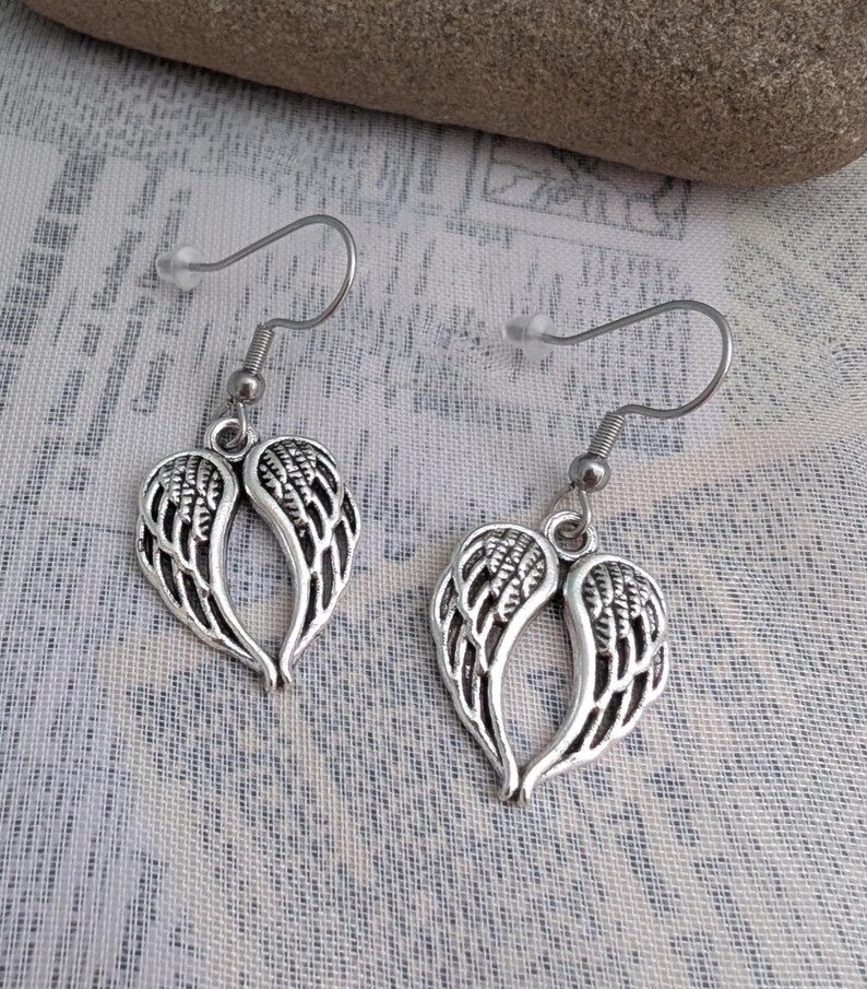 May include: A pair of silver-colored earrings featuring a wing design. Each earring has a hook for pierced ears and a detailed feather pattern within the wing shape. The earrings are displayed against a textured, light-colored background.