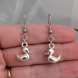 Peut inclure: Une paire de boucles d'oreilles pendantes en métal argenté représentant un petit canard en trois dimensions suspendu à un crochet d'oreille standard. Les breloques sont polies avec une surface lisse et réfléchissante, détaillées par un bec et un œil.