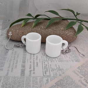 Peut inclure: Une paire de boucles d'oreilles en forme de tasses blanches en céramique. Chaque tasse miniature a une anse et est attachée à un crochet d'oreille argenté. Les boucles d'oreilles sont présentées sur une pierre avec du feuillage vert.