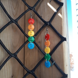 May include: A pair of rainbow button earrings. Each earring features six buttons in a rainbow pattern, starting with red and ending with blue. The earrings are suspended from silver hooks.