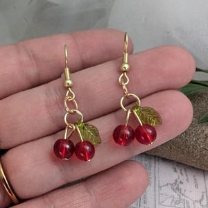 May include: A pair of gold-toned dangle earrings featuring red cherry beads and small green leaf accents. The earrings are designed to resemble a cluster of cherries, with a delicate and playful aesthetic.