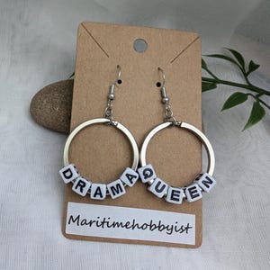 May include: Silver hoop earrings with white letter beads spelling "Drama Queen".