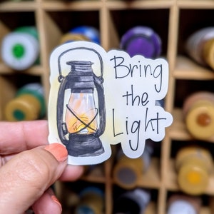 May include: A white sticker with a hand-drawn illustration of a black oil lamp with a yellow flame. The text "Bring the Light" is written in black cursive script.