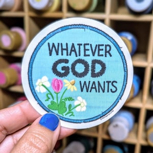 May include: A round blue and white embroidered patch with the text "Whatever God Wants" in black embroidery. The patch has a white border and a blue inner circle with white flowers and green leaves.
