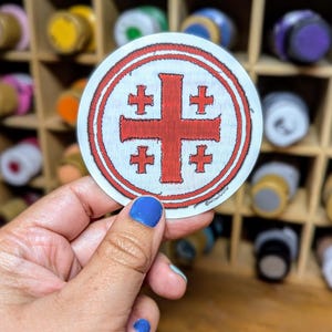 May include: A white round sticker with a red cross and four smaller red crosses inside a double circle. The sticker is being held by a hand with blue nail polish.