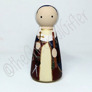 May include: Hand-painted wooden peg doll depicting a saint. The doll is wearing a blue and red robe with gold accents. The saint is holding a cross and a staff.
