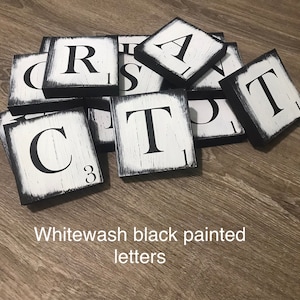 Op de afbeelding: Witgewassen zwart geschilderde letters op vierkante houten blokken. De letters zijn zwart, met een verweerde witte achtergrond en zwarte randen. De tekst "Whitewash black painted letters" staat onderaan de afbeelding.