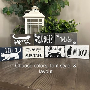 May include: Six wooden blocks with cat names and paw prints painted on them. The names are Bella, Fluffy, Boots, Milo, Seth, Blacky, and Willow. The blocks are arranged in two rows. The text below the blocks reads "Choose colors, font style, & layout".