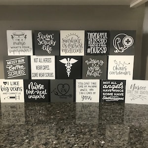 Può includere: Una collezione di 15 piccoli cartelli in legno con testo bianco e nero. I cartelli presentano vari detti e frasi relative all'infermieristica, tra cui "Sono un'infermiera, qual è il tuo superpotere?", "La luce del sole è la migliore medicina", "Le infermiere danno il ritmo" e "Non tutti gli angeli hanno le ali, alcuni hanno stetoscopi".