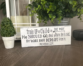 Puede incluir: Un letrero de madera blanco con texto negro que dice "Instruye al niño en el camino en que debe andar, y aun cuando fuere viejo no se apartará de él. Proverbios 22:6".