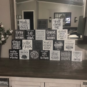 May include: A collection of square wooden blocks arranged in a pyramid, each featuring black or white painted text with various kitchen and coffee-themed phrases such as Made with Love, King of the Kitchen, and All You Need is Coffee.