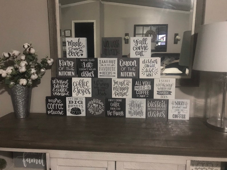 May include: A collection of square wooden blocks arranged in a pyramid, each featuring black or white painted text with various kitchen and coffee-themed phrases such as Made with Love, King of the Kitchen, and All You Need is Coffee.