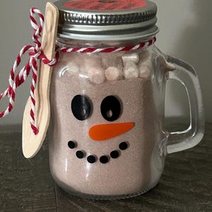 May include: A clear glass mason jar mug filled with brown cocoa powder and white marshmallows. The front features a snowman face made of black and orange decals. A wooden spoon is tied to the handle with red and white twine, and a metal lid seals the top.