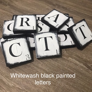 Op de afbeelding: Wit gewassen zwart geschilderde letters. Vierkante houten tegels met zwarte randen en witte, verweerde oppervlakken. Elke tegel heeft een grote zwarte letter. De tekst in de afbeelding luidt: Wit gewassen zwart geschilderde letters.