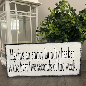 Op de afbeelding: Een rechthoekig houten bord met de tekst "Having an empty laundry basket is the best five seconds of the week." Het bord heeft een witte achtergrond met zwarte letters en een donkergrijze rand. Een witte lantaarn en groene plant staan op de achtergrond.