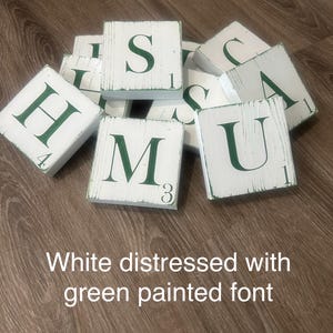 Op de afbeelding: Witte, verweerde houten lettertegels met groen geschilderde letters en cijfers. De letters zijn H, S, M, U en A. De tekst in de afbeelding luidt: "Wit verweerd met groen geschilderd lettertype."