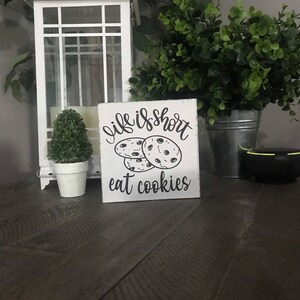 May include: A square white wooden sign with distressed edges featuring black text that reads "life is short" above a graphic of three chocolate chip cookies, with the text "eat cookies" printed in black lettering at the bottom of the sign.