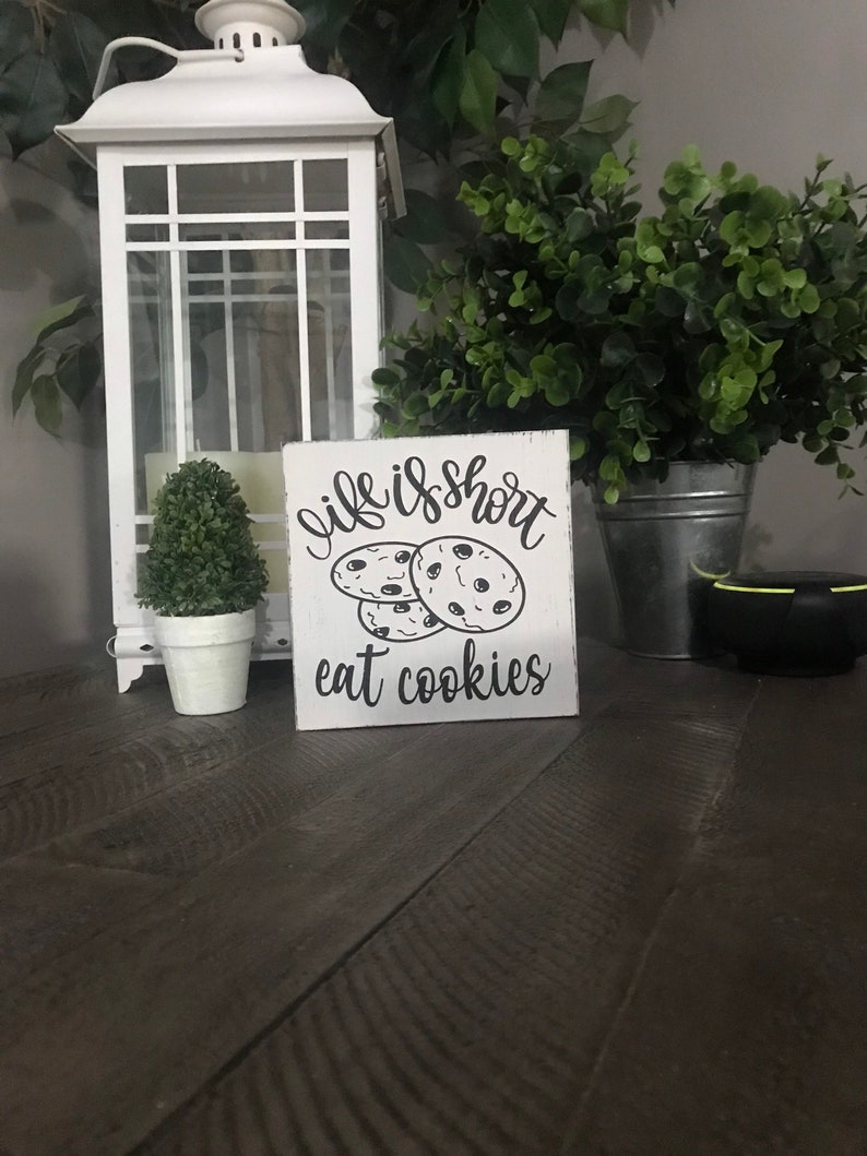 May include: A square white wooden sign with distressed edges featuring black text that reads "life is short" above a graphic of three chocolate chip cookies, with the text "eat cookies" printed in black lettering at the bottom of the sign.