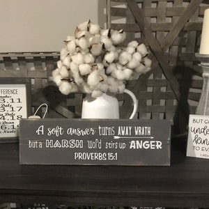 May include: A wooden sign with a white background and black text. The sign reads "A soft answer turns away wrath but a harsh word stirs up anger. Proverbs 15:1".