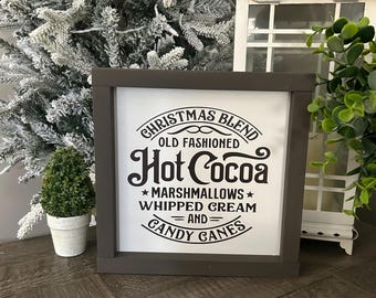 Op de afbeelding: Een ingelijst bord met de tekst "CHRISTMAS BLEND, OLD FASHIONED Hot Cocoa, *MARSHMALLOWS* WHIPPED CREAM =AND= CANDY CANES" in zwarte letters. Het frame is donkergrijs. Een kleine pot met een plant en een witte lantaarn staan op de achtergrond.