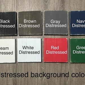 Op de afbeelding: Acht vierkante houten blokken met een distressed afwerking, elk in een andere kleur: zwart, bruin, grijs, marineblauw, cr&egrave;me, wit, rood en groen. Elk blok is voorzien van de kleurnaam en het woord "Distressed". De tekst "Distressed background colors" staat eronder.