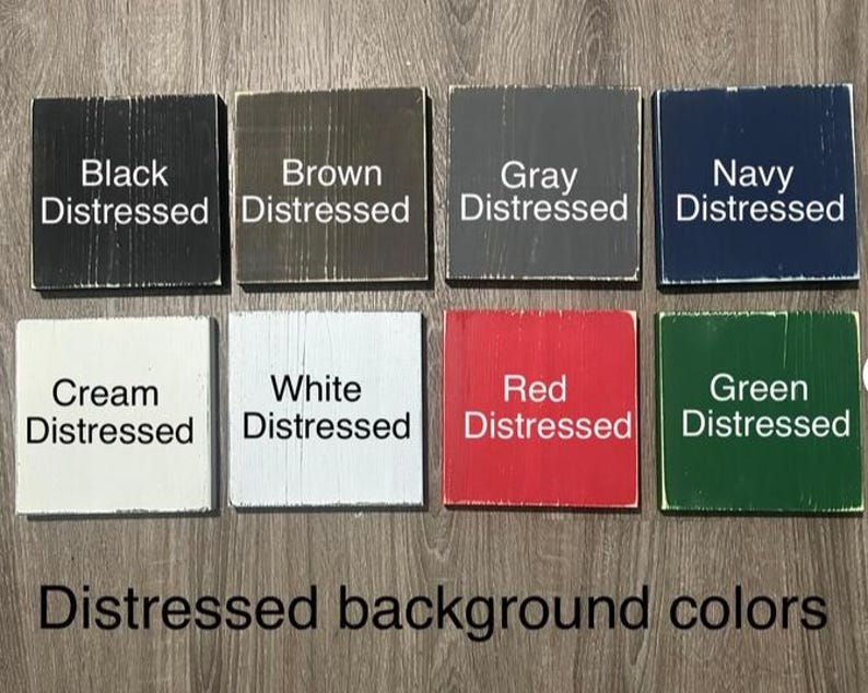 Op de afbeelding: Acht vierkante houten blokken met een distressed afwerking, elk in een andere kleur: zwart, bruin, grijs, marineblauw, cr&egrave;me, wit, rood en groen. Elk blok is voorzien van de kleurnaam en het woord "Distressed". De tekst "Distressed background colors" staat eronder.