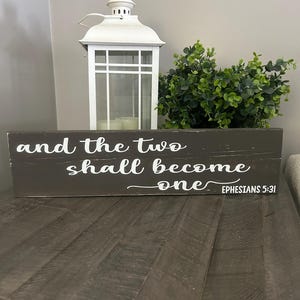 May include: A wooden sign with the text "and the two shall become one - Ephesians 5:31" in white lettering. The sign is sitting on a wooden table with a white lantern and a green plant in the background.