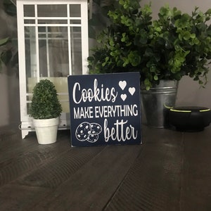May include: A square navy blue wooden sign with white text that reads "Cookies MAKE EVERYTHING better" accompanied by white heart icons and a graphic illustration of two chocolate chip cookies positioned at the bottom left corner of the sign.