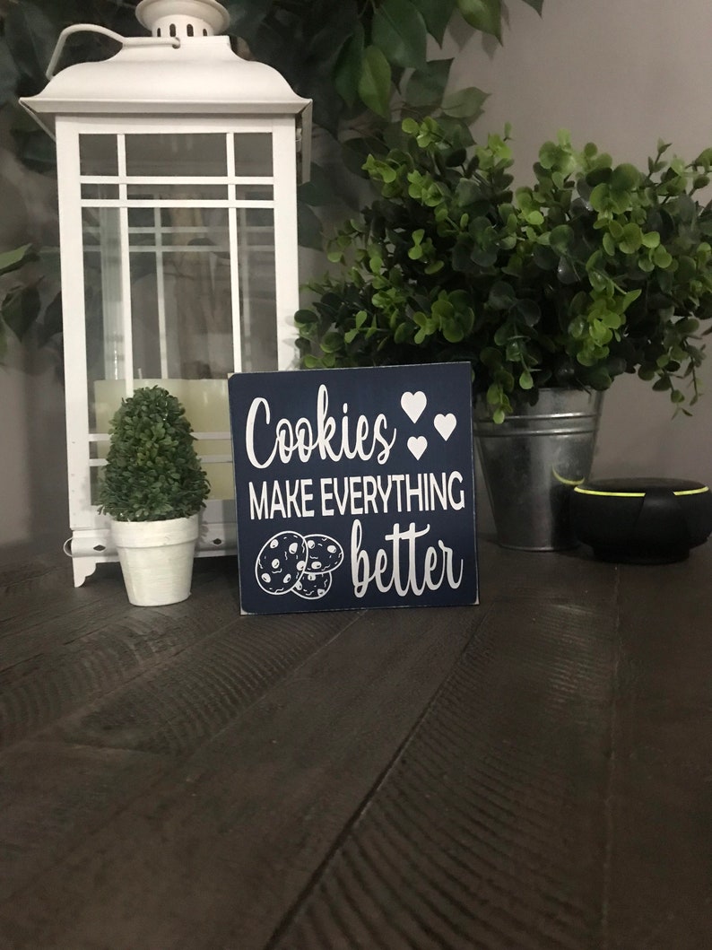 May include: A square navy blue wooden sign with white text that reads "Cookies MAKE EVERYTHING better" accompanied by white heart icons and a graphic illustration of two chocolate chip cookies positioned at the bottom left corner of the sign.