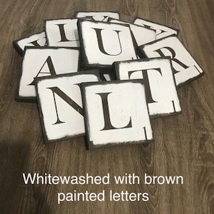 Op de afbeelding: Witgekalkte houten lettertegels met bruin geschilderde randen en zwarte letters. De tekst "Whitewashed with brown painted letters" staat onderaan. De tegels liggen op een houten oppervlak.