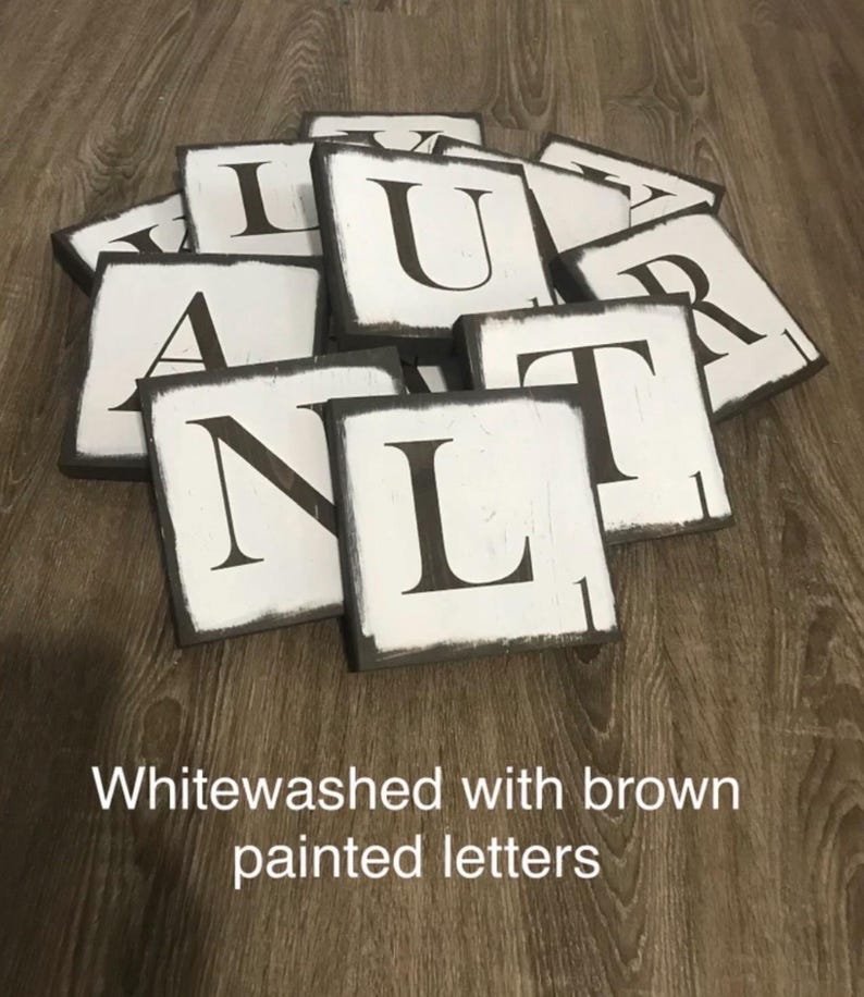 Op de afbeelding: Witgekalkte houten lettertegels met bruin geschilderde randen en zwarte letters. De tekst "Whitewashed with brown painted letters" staat onderaan. De tegels liggen op een houten oppervlak.
