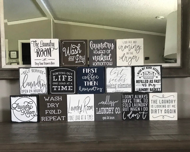 May include: A collection of square wooden signs with various laundry-themed quotes in black and white lettering. The signs are arranged on a dark surface, with phrases like "Laundry Room," "Wash and Dry," and "First Coffee Then Laundry."