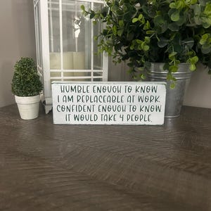 以下が含まれることがあります： 「HUMBLE ENOUGH TO KNOW I AM REPLACEABLE AT WORK. CONFIDENT ENOUGH TO KNOW IT WOULD TAKE 4 PEOPLE」と書かれた黒い文字の白い木製サイン。サインは木製の表面にあり、白いランタンと緑の植物が背景にあります。