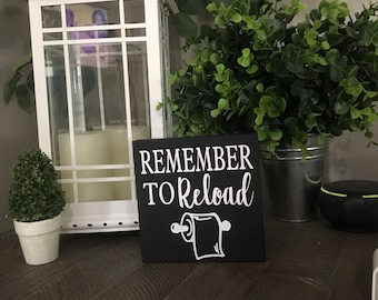 Peut inclure: Une pancarte en bois noir avec du texte blanc qui dit "Remember to Reload" avec une illustration blanche d'un rouleau de papier toilette.