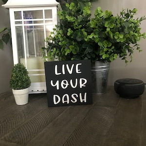 May include: Black wooden sign with white text that reads "Live Your Dash". The sign is sitting on a wooden table with a small potted plant and a white lantern in the background.