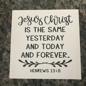 May include: A square white sign featuring black text that reads Jesus Christ is the same yesterday and today and forever. Below the text is a decorative black leaf motif, followed by the citation Hebrews 13:8 in a simple sans-serif font.
