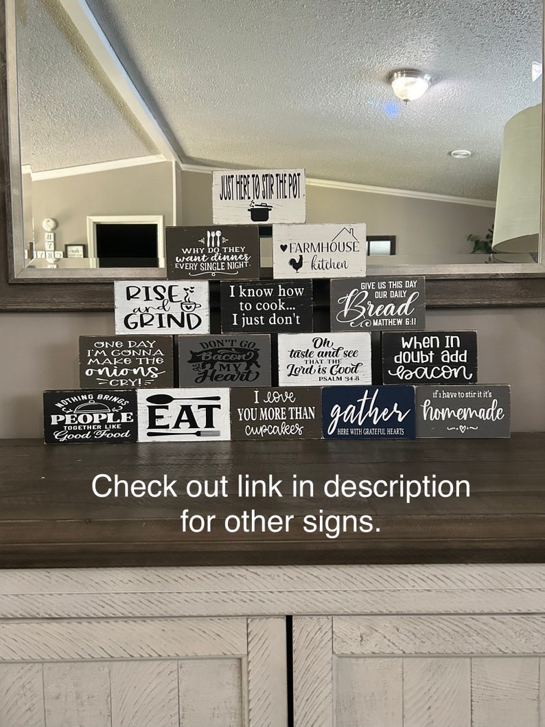 May include: A collection of rectangular wooden signs stacked in a pyramid shape. Each sign features black or white painted backgrounds with contrasting text and graphics related to cooking, food, and kitchen themes, including phrases like Rise and Grind, Farmhouse Kitchen, and Eat.