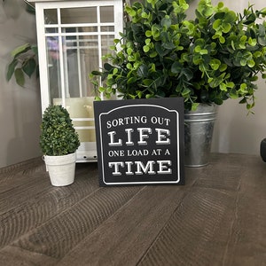 May include: Black wooden sign with white text that reads "Sorting out life one load at a time". The sign is sitting on a wooden table with a white lantern and two small potted plants.