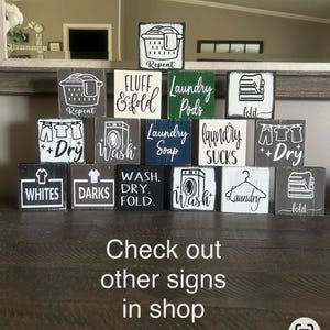 May include: A collection of small, square wooden signs with laundry-themed designs and text. The signs feature images of laundry baskets, washing machines, and clothing, with phrases like "Wash," "Dry," and "Fold." The text "Check out other signs in shop" is also visible.