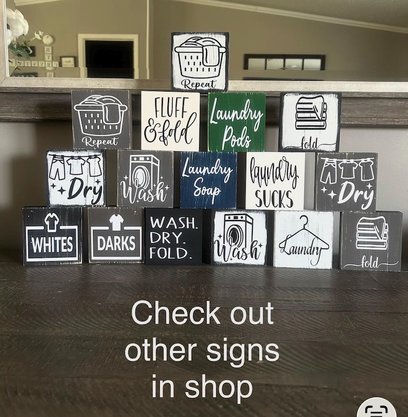 May include: A collection of small, square wooden signs with laundry-themed designs and text. The signs feature images of laundry baskets, washing machines, and clothing, with phrases like "Wash," "Dry," and "Fold." The text "Check out other signs in shop" is also visible.