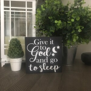 May include: A black square sign with white text reading "Give it to God and go to sleep." The sign features a crescent moon and star. A white lantern, potted plant, and greenery are in the background.