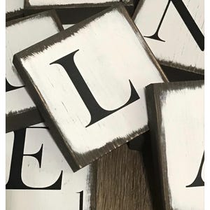 Op de afbeelding: Close-up van verschillende vierkante houten blokken met wit geschilderde oppervlakken en zwarte letters. De blokken hebben een verweerde, witgekalkte bruine rand. De tekst "Whitewash brown with black font" staat onderaan.