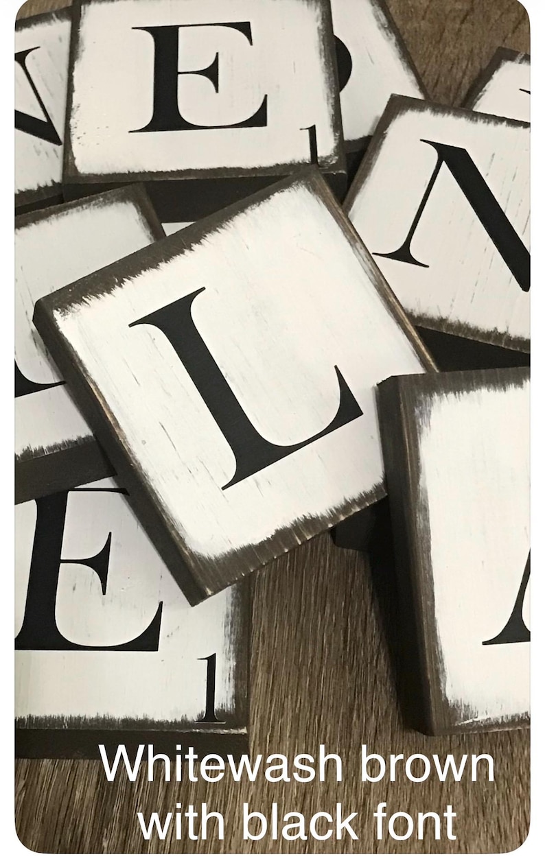 Op de afbeelding: Close-up van verschillende vierkante houten blokken met wit geschilderde oppervlakken en zwarte letters. De blokken hebben een verweerde, witgekalkte bruine rand. De tekst "Whitewash brown with black font" staat onderaan.