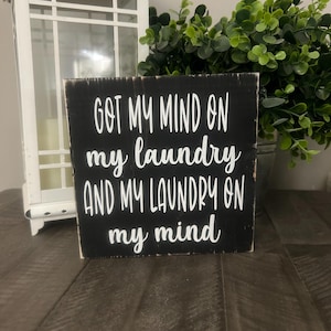 May include: A black wooden sign with white lettering that reads "GOT MY MIND ON my laundry AND MY LAUNDRY ON my mind." A white lantern and greenery are in the background.