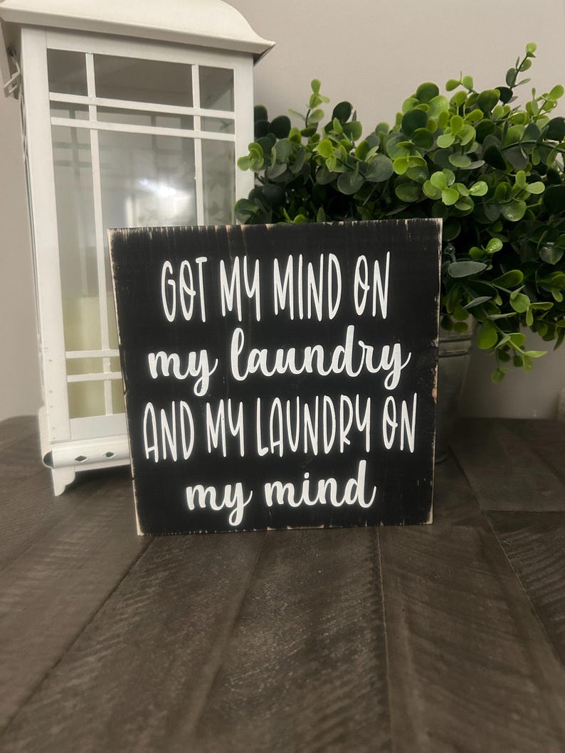 May include: A black wooden sign with white lettering that reads "GOT MY MIND ON my laundry AND MY LAUNDRY ON my mind." A white lantern and greenery are in the background.
